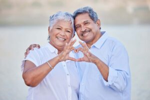 A happy senior couple showing a heart shape with their fingers and enjoying fresh air on vacation at the beach while bonding. Portrait of retired couple hugging at beach with smile and love together
